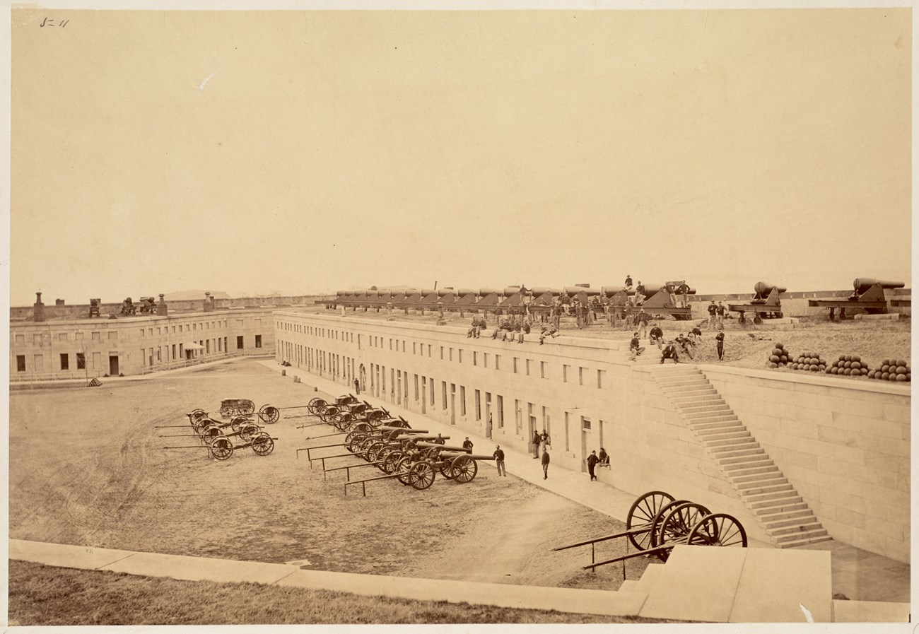 Sepia photo of the interior of Fort Warren with soldiers on the top of the fort and guns lined up on the grounds.
