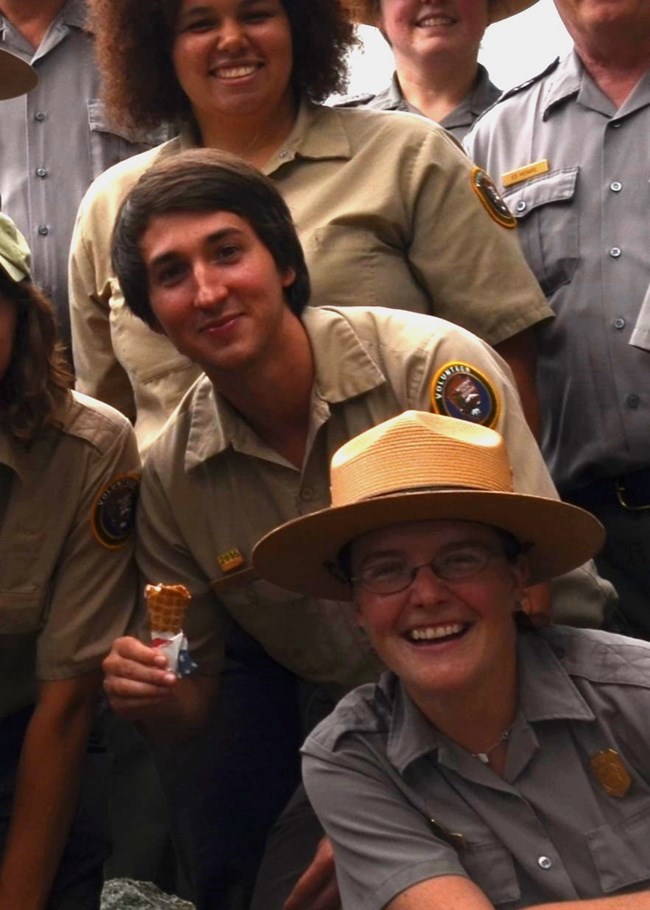 A young man with brown hair holds a half-eaten ice cream cone wearing a tan NPS volunteer shirt and is surrounded by smiling faces in NPS uniforms.