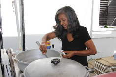 Isabel Pizarro Woman with black and grey shoulder-length hair stands in an outdoor kitchen stirring a large pot of rice.