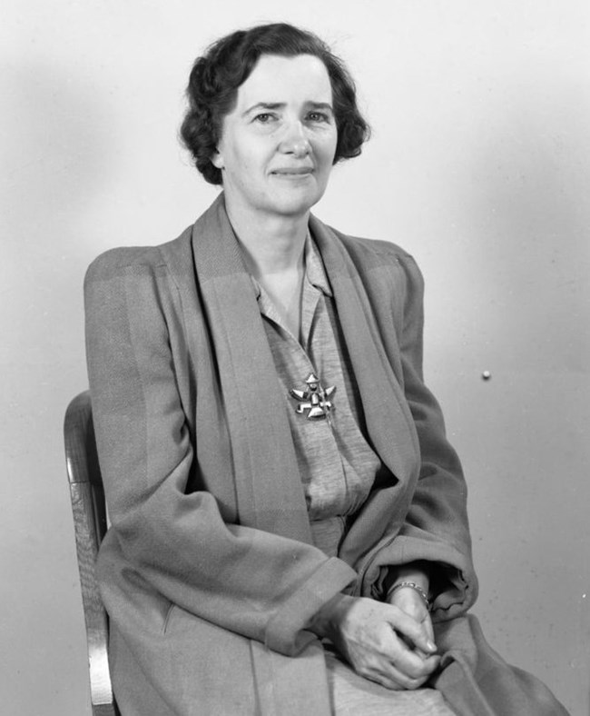 Isabel Story sits in a chair in front of a plain background wearing a dress and coat.