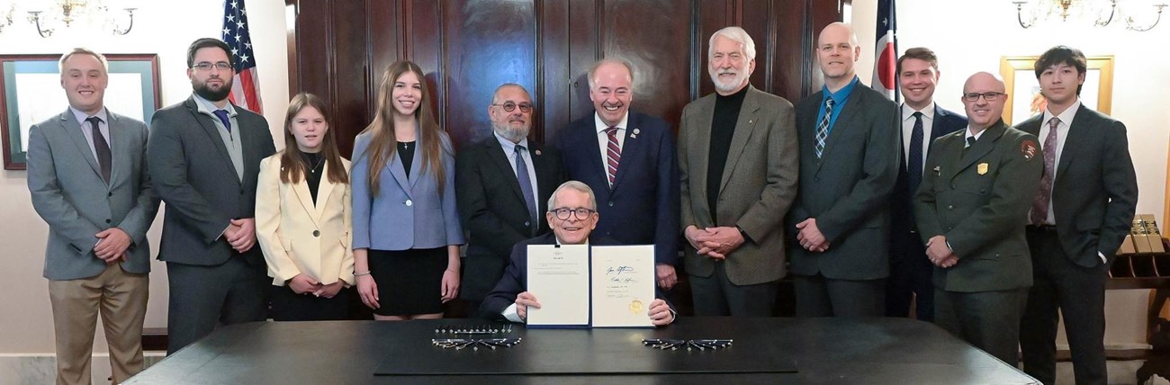 Ohio Governor DeWine is joined by James A. Garfield's descendants and other dignitaries at the bill signing to designate November 19 as James A. Garfield Day in Ohio. A group of people stand behind a table that a man sits at, holding a sheaf of papers.