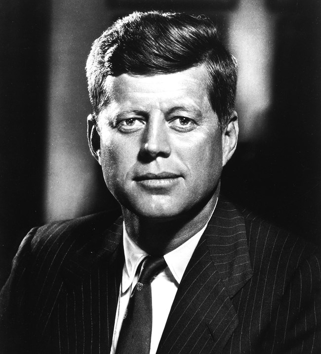 President Kennedy in a dark suit stares straight at the camera.