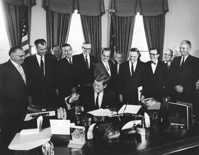 A group of government leaders stand behind President Kennedy.  Kennedy is signing a document and smiling.