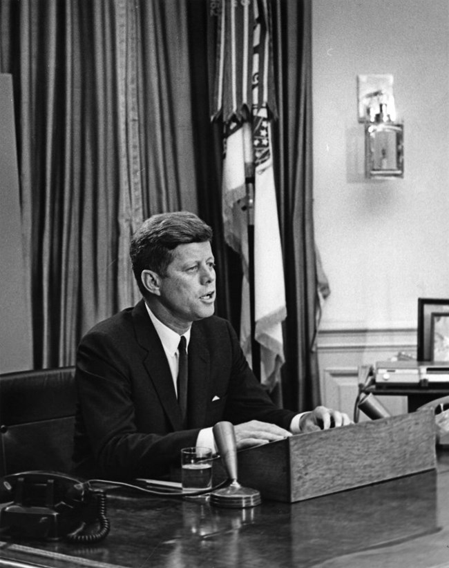 President John F. Kennedy delivers an address seated at the resolute desk in the oval office.