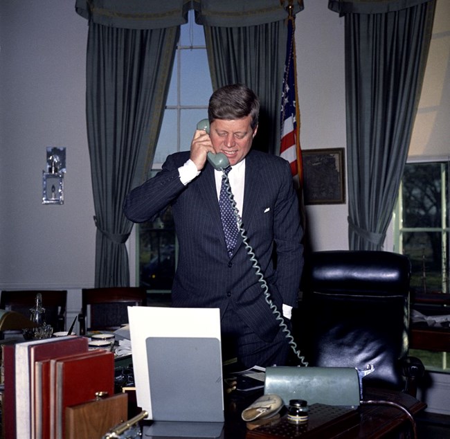 President Kennedy standing behind his desk in the Oval Office talking on the phone.