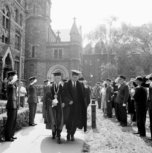 President Kennedy walks with a group of men in graduation regalia