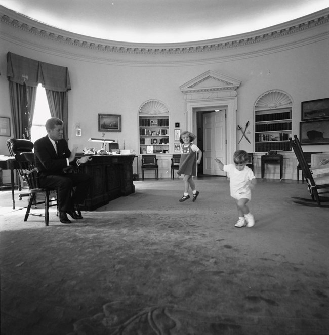 JFK seated in a chair near his desk in the Oval Office claps as his children, Caroline and John Jr. dance.