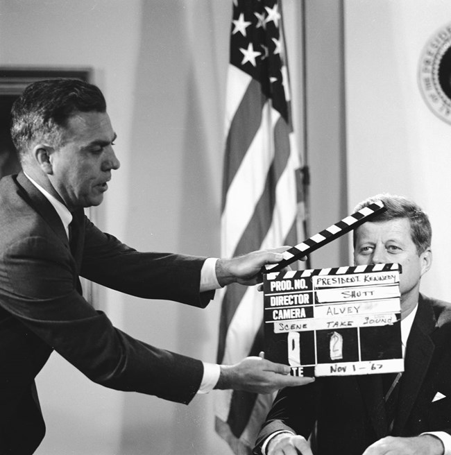 John F. Kennedy is seated.  A man with a film clapboard conceals part of Kennedy's face as the film is about to start.