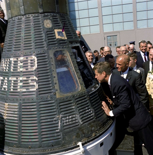 John F. Kennedy and John Glenn, wearing suits, look into space capsule, Friendship 7.  The capsule is on the left and there is a crowd of onlookers behind them.