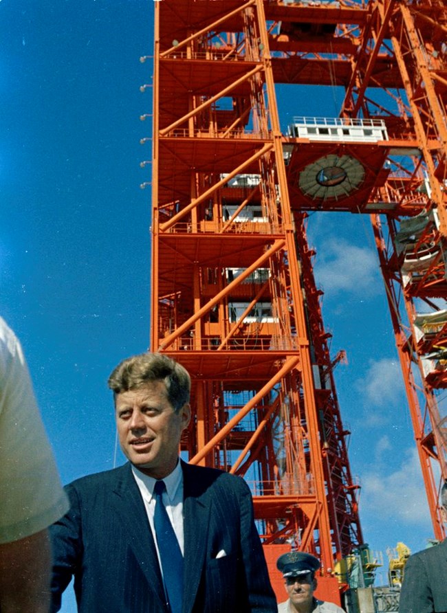 President Kennedy at a Saturn rocket launch site Kennedy is shown with towering red scaffolding behind him