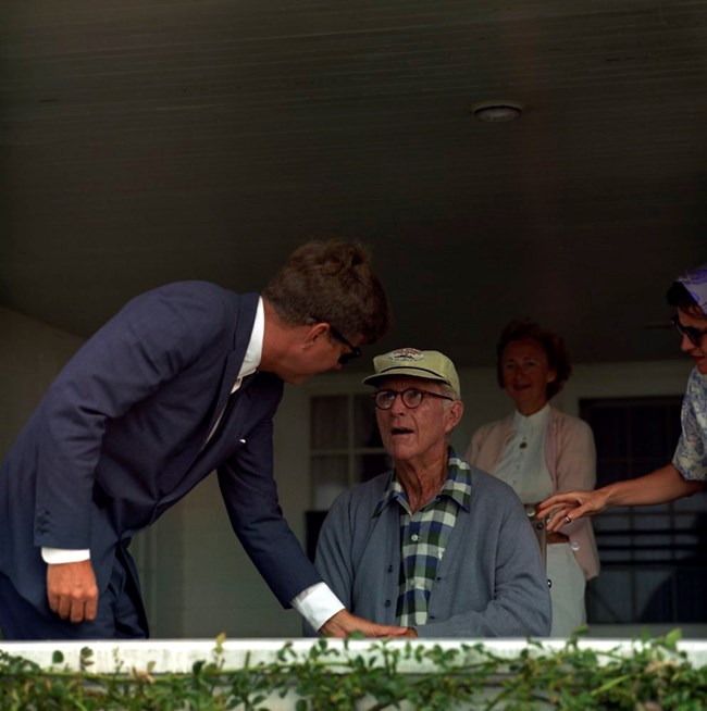 President Kennedy leans over to touch his father's hand.