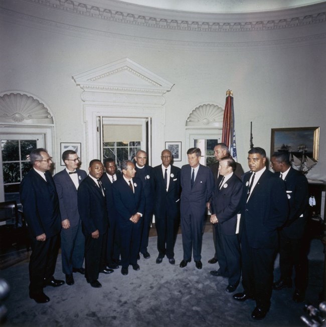 A group of men in suits stand in the Oval Office. A flag and portraits along the walls can be seen in the background.