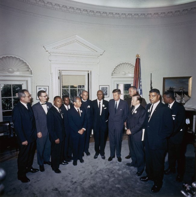 President Kennedy meets with organizers of "The March on Washington for Jobs and Freedom” A group of men in suits stand in the Oval Office. A flag and portraits along the walls can be seen in the background.