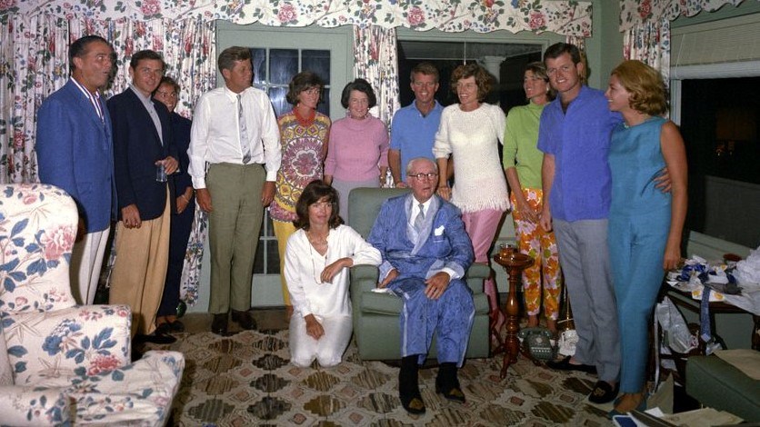 Kennedy family stands around Joe Kennedy Sr.. JPK Sr. sits in a chair wearing blue pajamas and a tie.