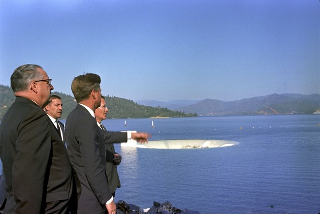 President Kennedy and a group of officials stand at the left of the photo.  They are standing near a water body.