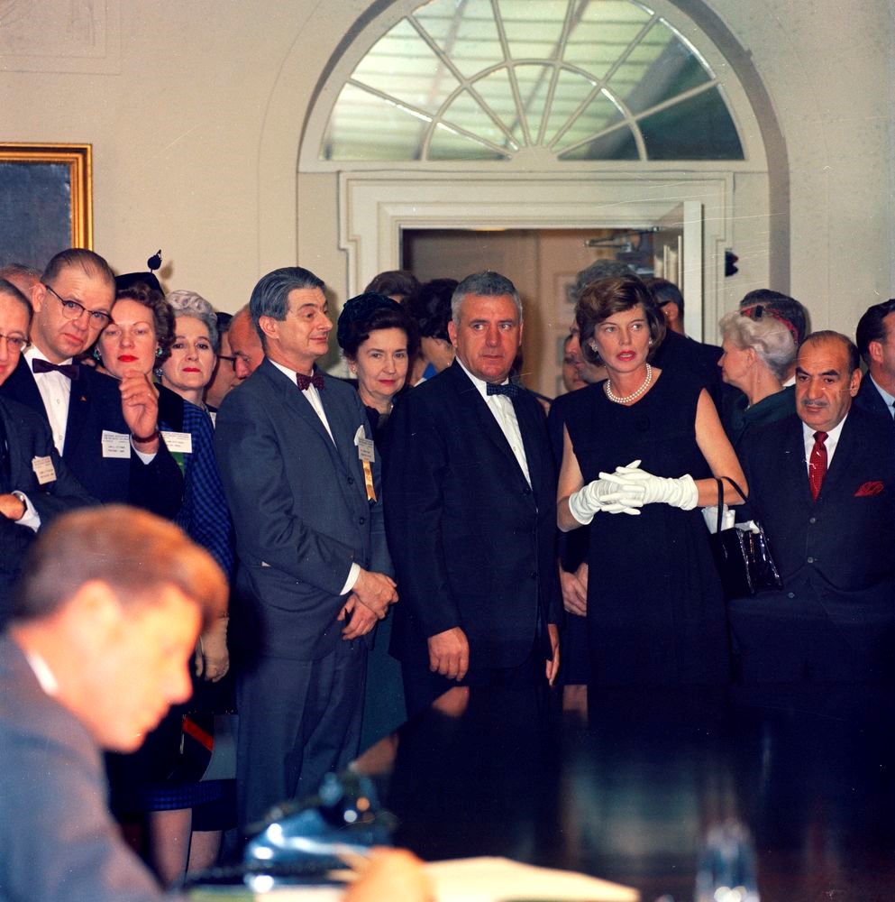 President Kennedy (out of focus in foreground) signs a bill. Eunice Kennedy stands in a group of people behind him.