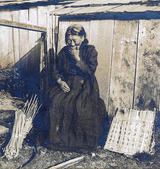 Indigenous woman sitting in front of wooden house in Euoropean garb. She is laughing into her hand. At her feet are plant materials for weaving
