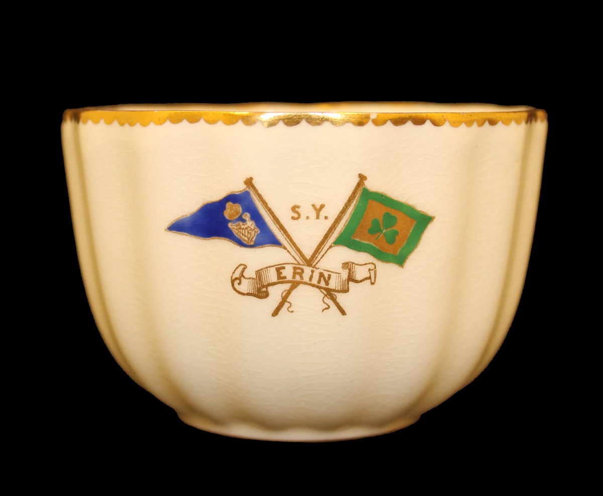 White teacup with image of two flags, one green with gold shamrock and one a blue pennant with harp