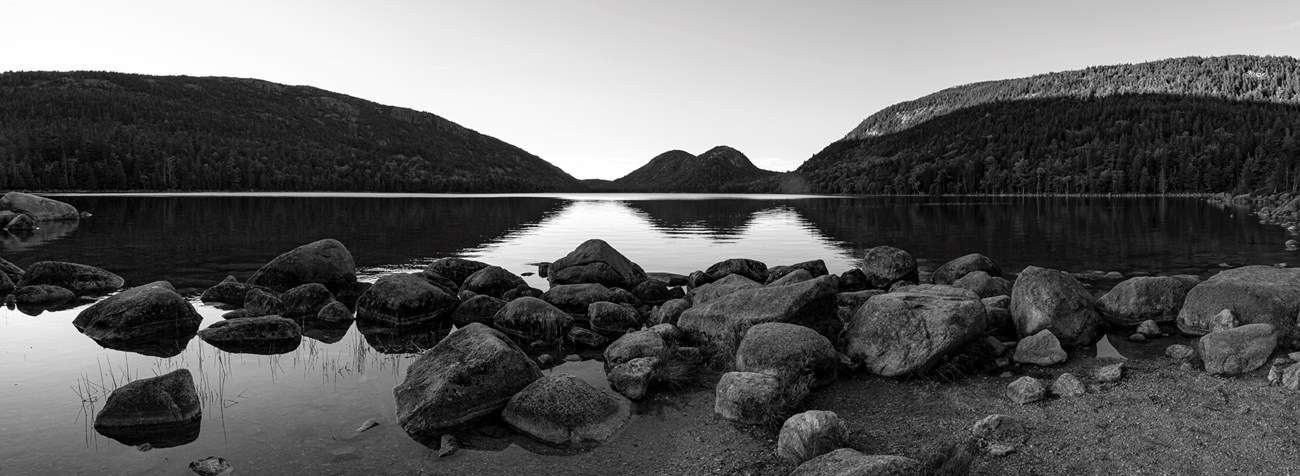 Black and white panoramic landscape photo with mountains in distance, a pond with ripple reflections, and boulders in the foreground Black and white panoramic landscape photo with mountains in distance, a pond with ripple reflections, and boulders in the foreground