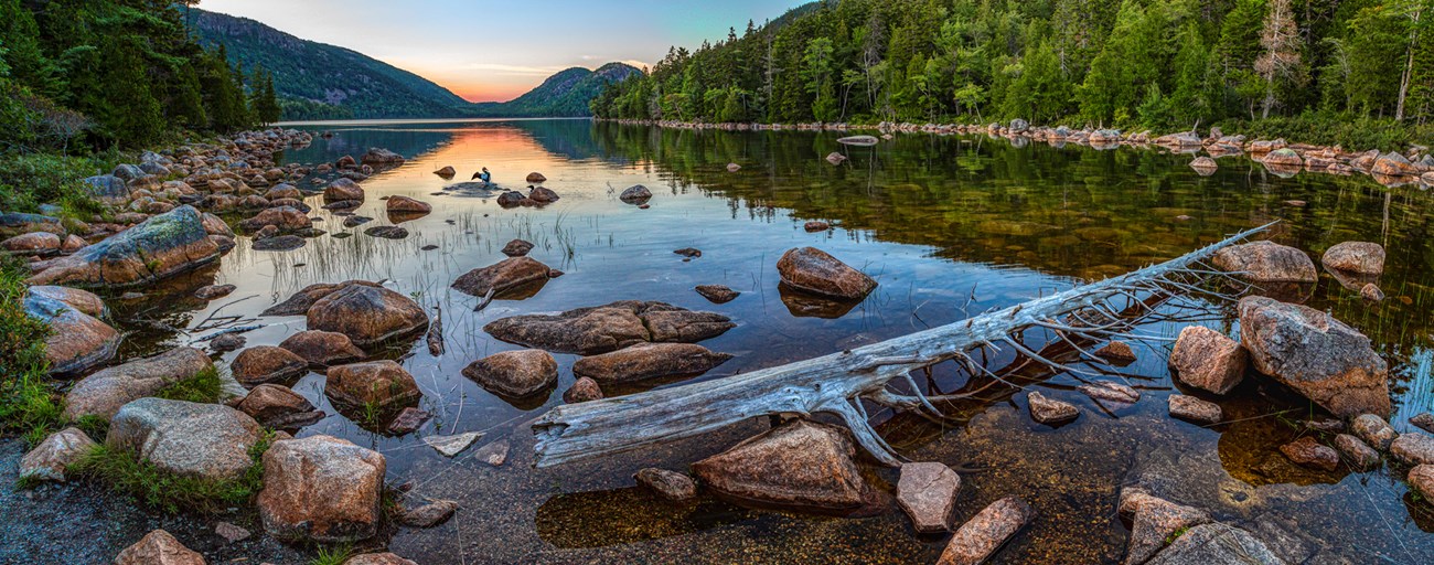 Landscape photo of a clear pond reflecting light with pink boulders and a weathered log in the foreground, trees lining the shoreline and mountains in the distance.