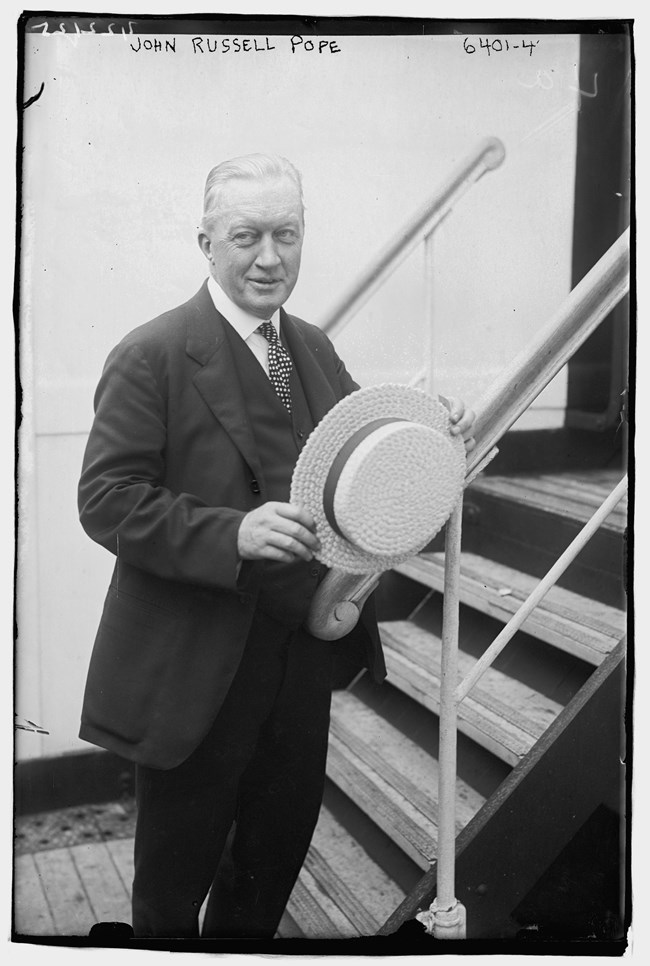 A man candidly posing in a suit and holding a flat-brimmed hat in front of his body. He is resting his left arm on a stair railing and appears to be mid-speech.