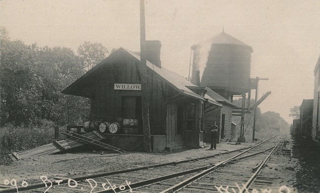 Black and white photo of a uniformed man standing outside the “Willow” train station. Barrels wait on the front platform. A water tower looms in the back. Text reads “090 B+O Depot, Willow, O.”