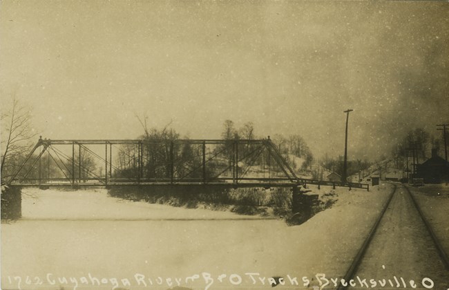 Station Rd. Bridge looking south ca 1907 Side view of a metal truss bridge over a frozen river blanketed in snow with railroad tracks and small buildings, right.