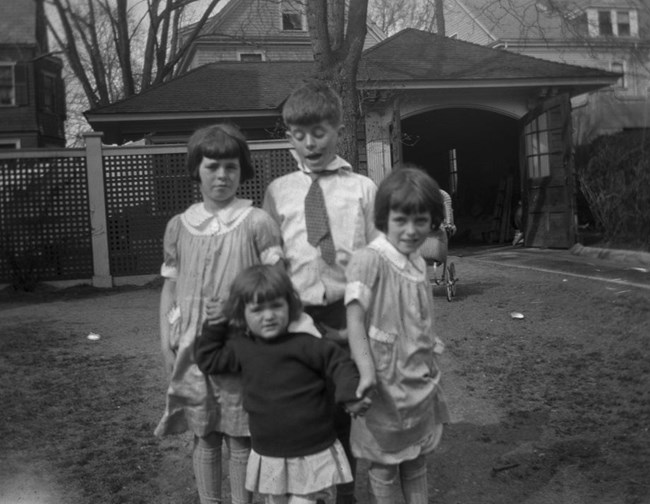 A black and white photo of four kids posing by a garage. A boy in a crooked collar blinks open mouthed behind two younger girls, in matching bob haircuts and collared dresses, who each hold one hand of a toddler in a skirt and sweater.