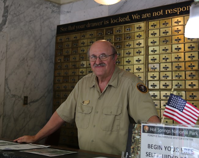A middle-aged white man with a mustache and glasses poses behind a front desk wearing an NPS volunteer uniform.