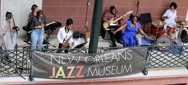 Jade Perdue singing into a microphone with a band of seven people on various instruments with a large banner in front that says, New Orleans Jazz Museum.