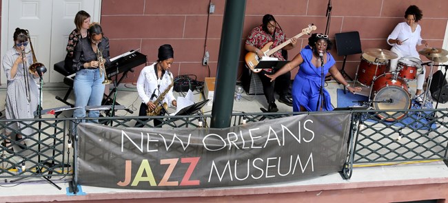 Jade Perdue. Jade Perdue singing into a microphone with a band of seven people on various instruments with a large banner in front that says, New Orleans Jazz Museum.