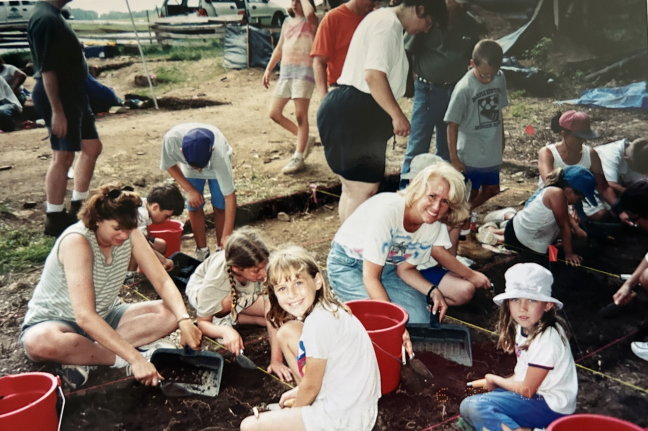 A group of children and adults working at a shallow archaeology site outside.