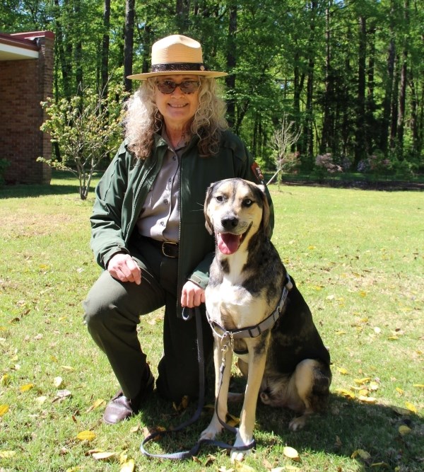 A woman in a park ranger uniform kneeling with her dog on grass.