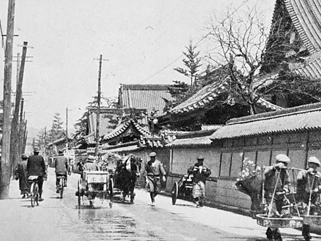Black and white photo of Hiroshima during World War II. Many people are biking and walking through a narrow street.