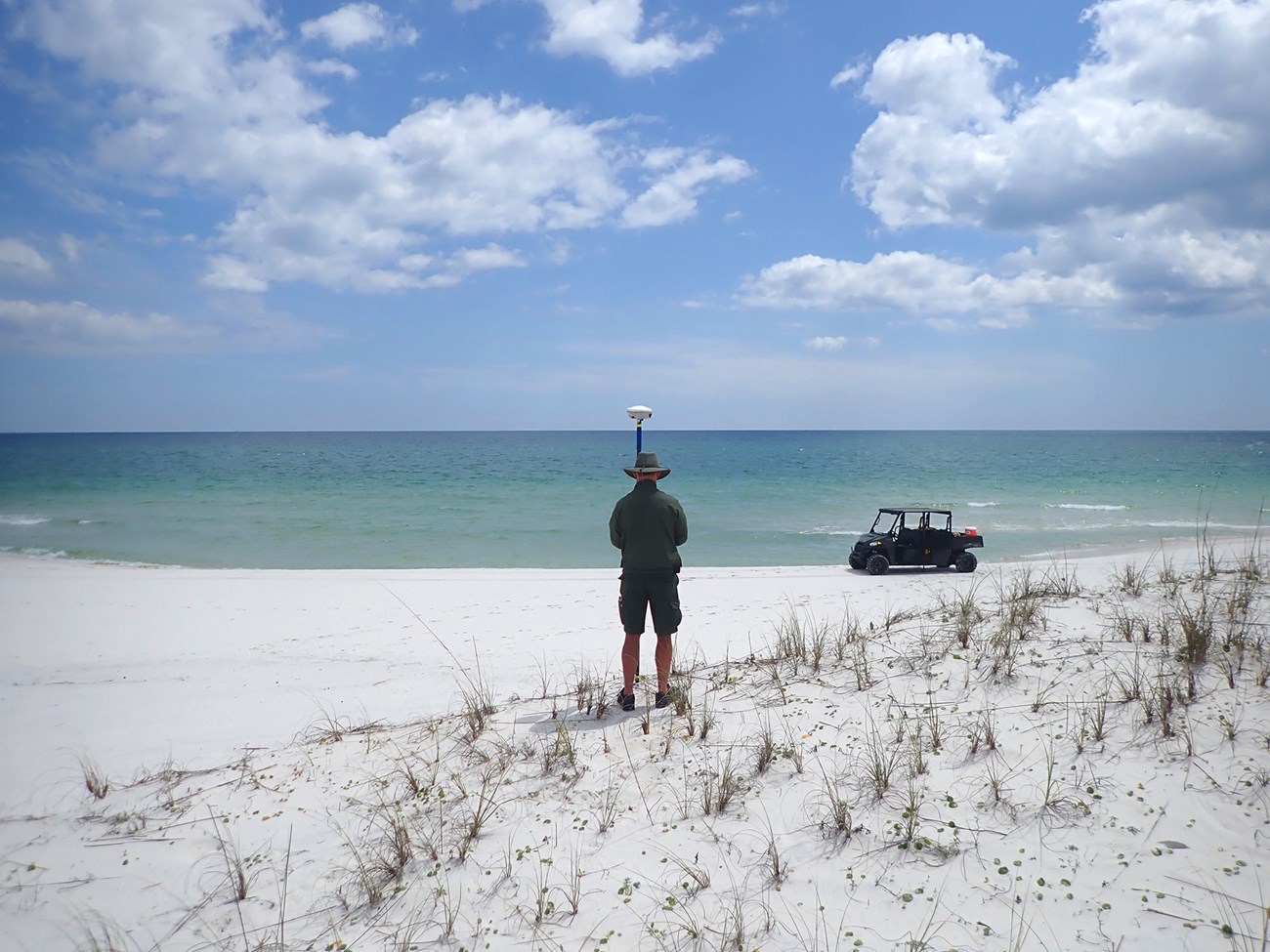 Man wearing an NPS uniform, stands with his back to the camera, holding a surveying instrument. The beach below him is white, with sparse beach grass. In front of him is an ATV with a turquoise ocean behind it.