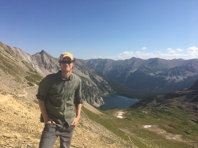 Man in hiking clothes, ball cap, and sunglasses poses in remote landscape with mountains and lake behind him.