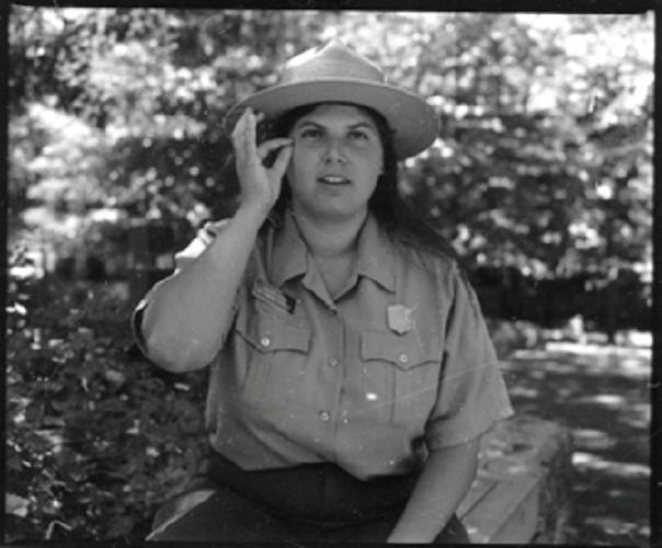 A black & white photo of Jennifer Jacobs, a white woman with straight dark hair past her shoulders. She sits outside in a park ranger uniform & hat. Her right hand is raised near her eye with index and thumb pinched and her other 3 fingers straight up.