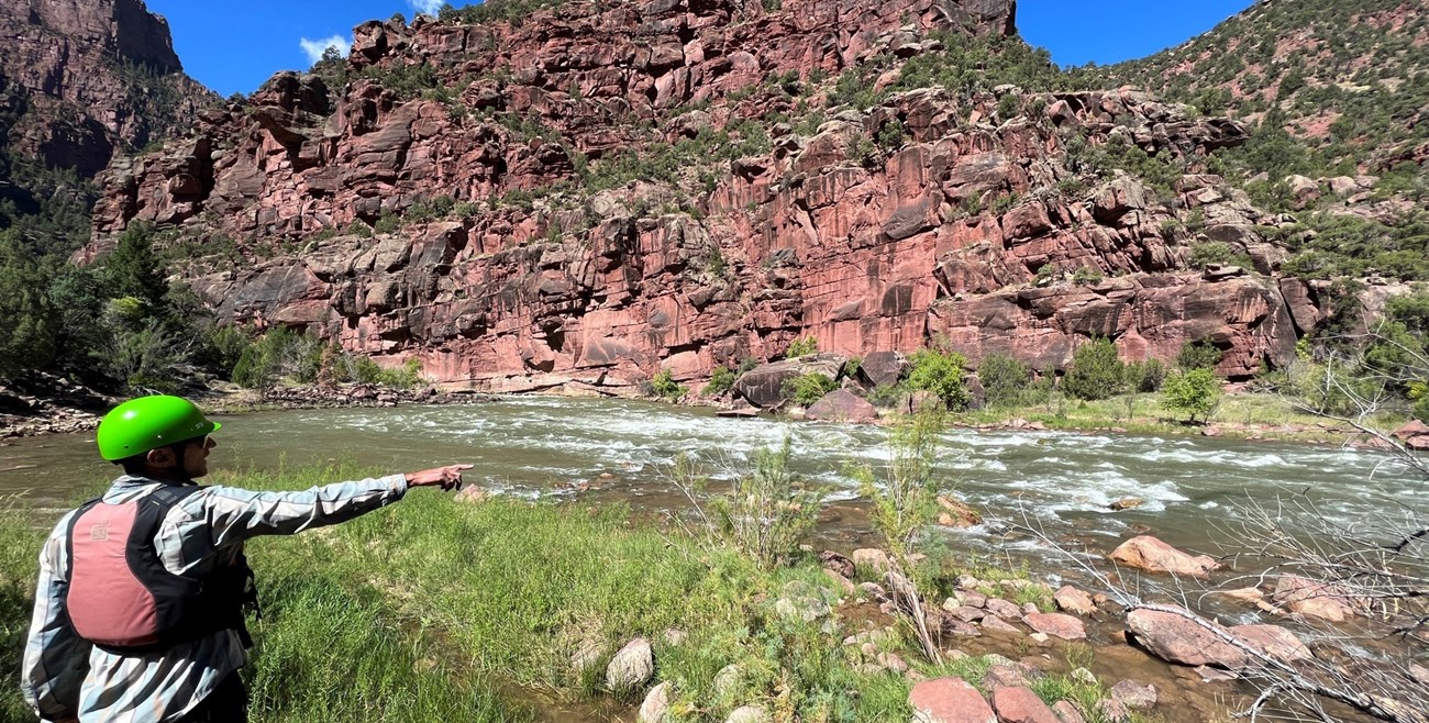 A man, wearing a helmet and a personal flotation device, pointing at a section of a whitewater rapid, with the canyon wall in the background.