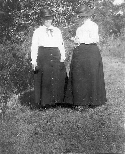 Two women wearing dark skirts, white blouses and dark hats stand facing us in front of a tree line.