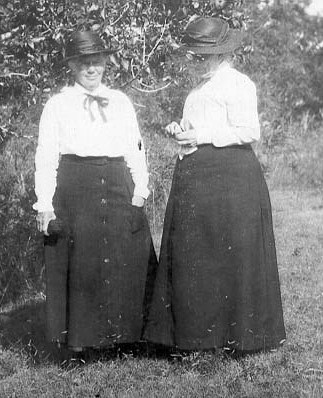 Jessie Foote Jack and friend Jessie Jack and Elizabeth Owen stand in a grassy patch in front of a bush. Both wear ankle-length black skirts, white long-sleeved blouses, and black hats.