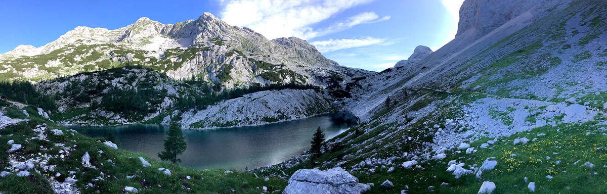 A panoramic image of a blue-green lake surrounded by rocky gray mountains, green meadows, green trees, and blue skies.