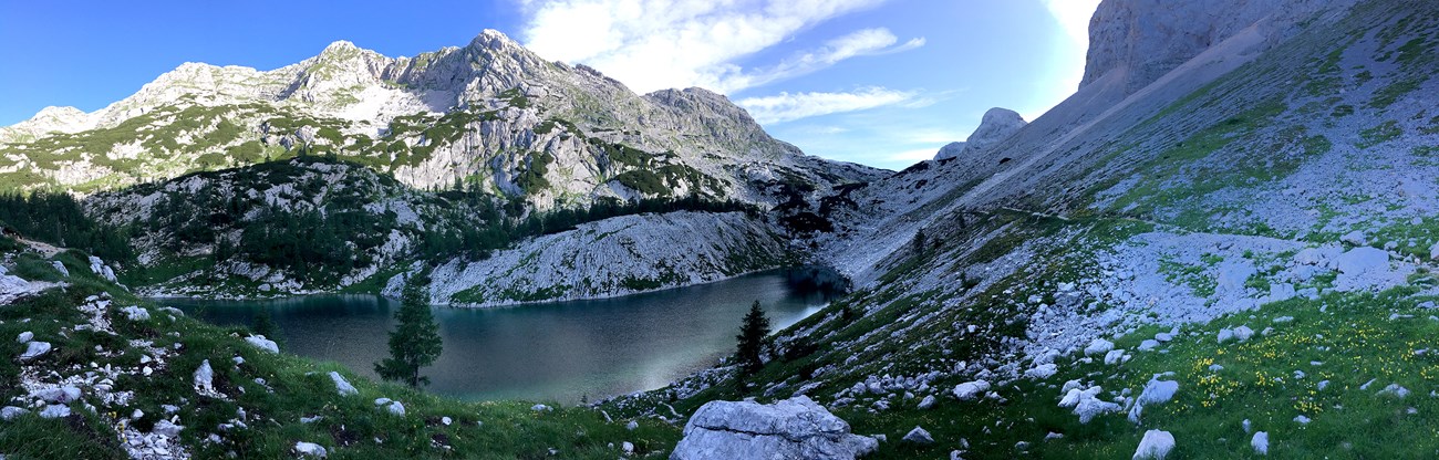 Jezero v Ledvicah, or "Lake at Ledvicah" A panoramic image of a blue-green lake surrounded by rocky gray mountains, green meadows, green trees, and blue skies.