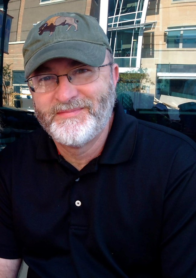 A middle-aged man with a salt and pepper beard and wearing glasses and a baseball cap