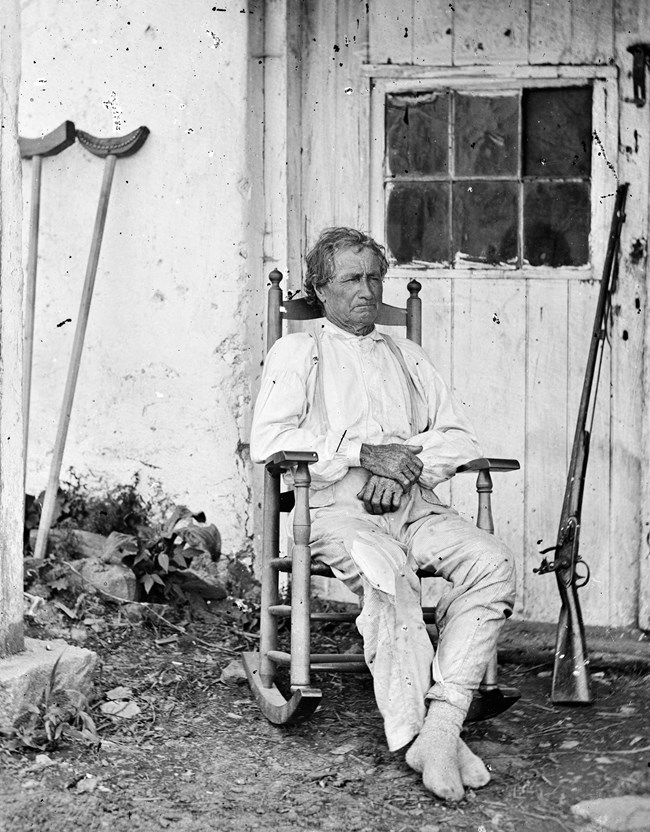 A man in his 60's sitting on a rocking chair with a gun and crutches near him.