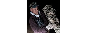 Action shot of John at night, wearing a headlamp, hat, binoculars, gloves, and maroon sweatshirt. He's lifting a metal mesh trap with a ferret inside.