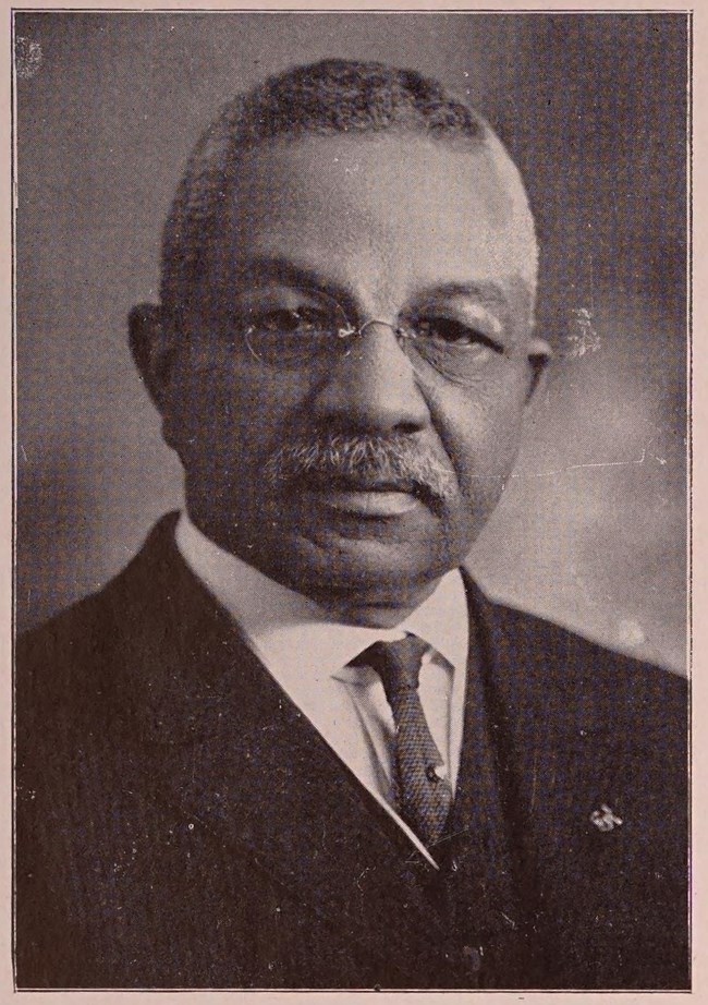 African American man with gray hair and a gray mustache wears glasses, a white shirt, dark tie, and a black suit.