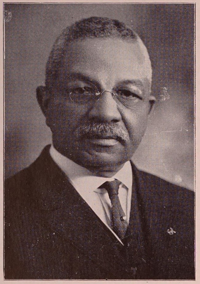John T. T. Warren African American man with gray hair and a gray mustache wears glasses, a white shirt, dark tie, and a black suit.