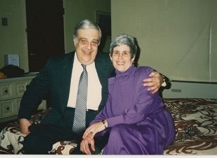 Man and woman seated on bed with a brown and white bedspread. The man has his left arm around the woman's shoulder. The man is wearing a dark suit, white shirt, and gray tie. The woman is wearing a purple dress.