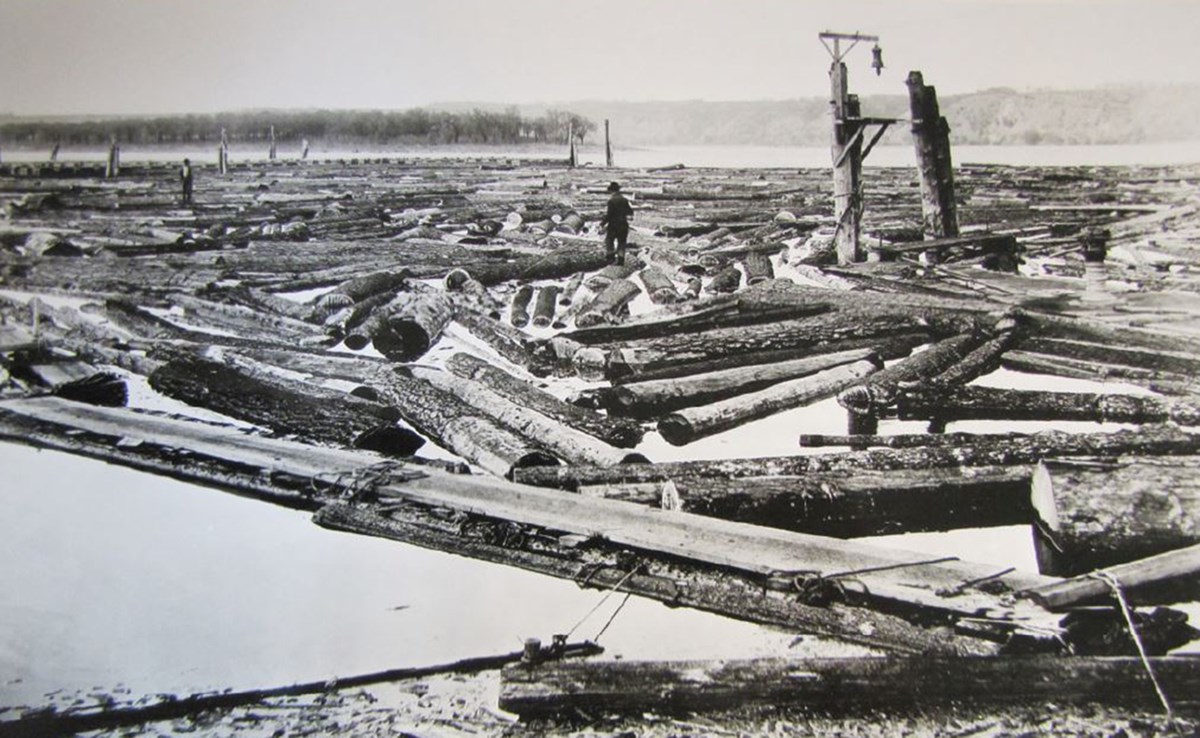 Black and white photo of logs on a river. Two men are standing on logs in the background.