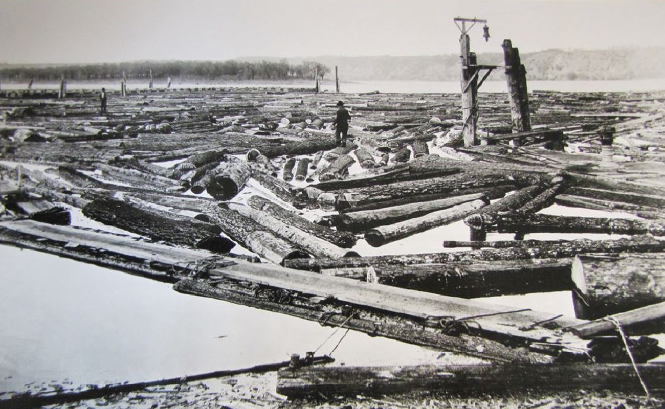 Black and white photo of logs on a river. Two men are standing on logs in the background.